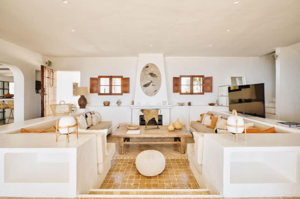 A spacious, modern living room with white walls and decor exudes a luxury travel vibe. It features two beige sofas with orange cushions facing a fireplace. A large TV is on the right, resting on tan tiles. A woven pouf in the center complements wooden accents to complete the look. David Pearson Travel