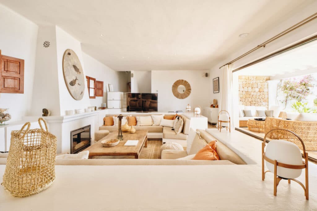 Spacious living room with white walls, a large beige sectional sofa, wicker chairs, and a wooden coffee table. Embracing the essence of luxury travel, a flat-screen TV is mounted on the wall, and a sliding glass door opens to a sunlit patio with additional seating and lush greenery. David Pearson Travel