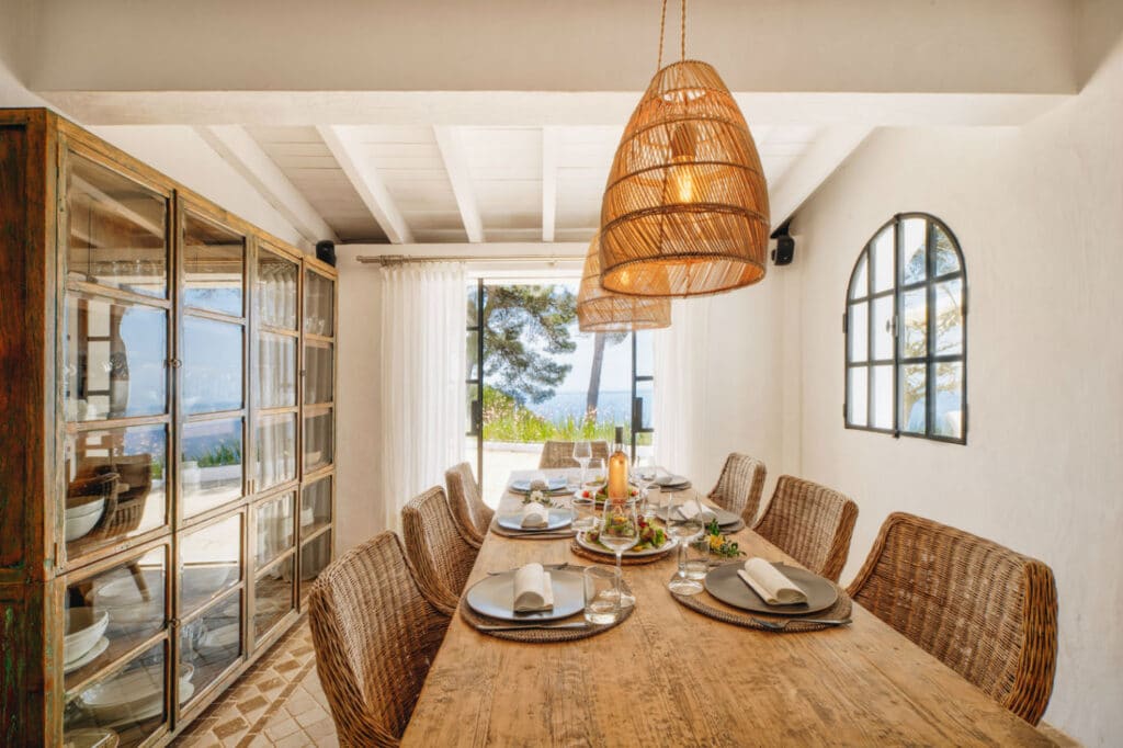 Dining room with a rustic wooden table set for eight, featuring wicker chairs and elegant tableware fit for luxury travel. Large wicker pendant lights hang from a white beamed ceiling, while a glass cabinet and windows offer breathtaking views of trees and the sea in the background. David Pearson Travel