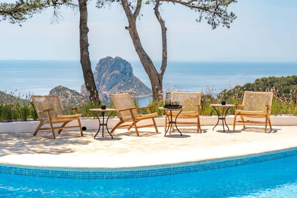 A serene poolside scene with wicker chairs and tables set on a patio exudes luxury travel vibes. Beyond, the view overlooks the sea and a large rocky island under a clear blue sky, framed by two tall trees. David Pearson Travel