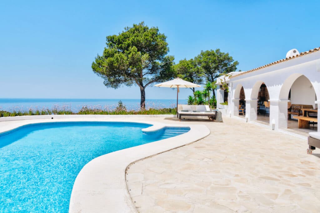 Experience the epitome of luxury travel at a Mediterranean villa with arched white walls and a serene pool. Two grand trees offer shade, against a backdrop of azure skies and ocean views. Relax under a beige umbrella in the elegant poolside seating area. David Pearson Travel