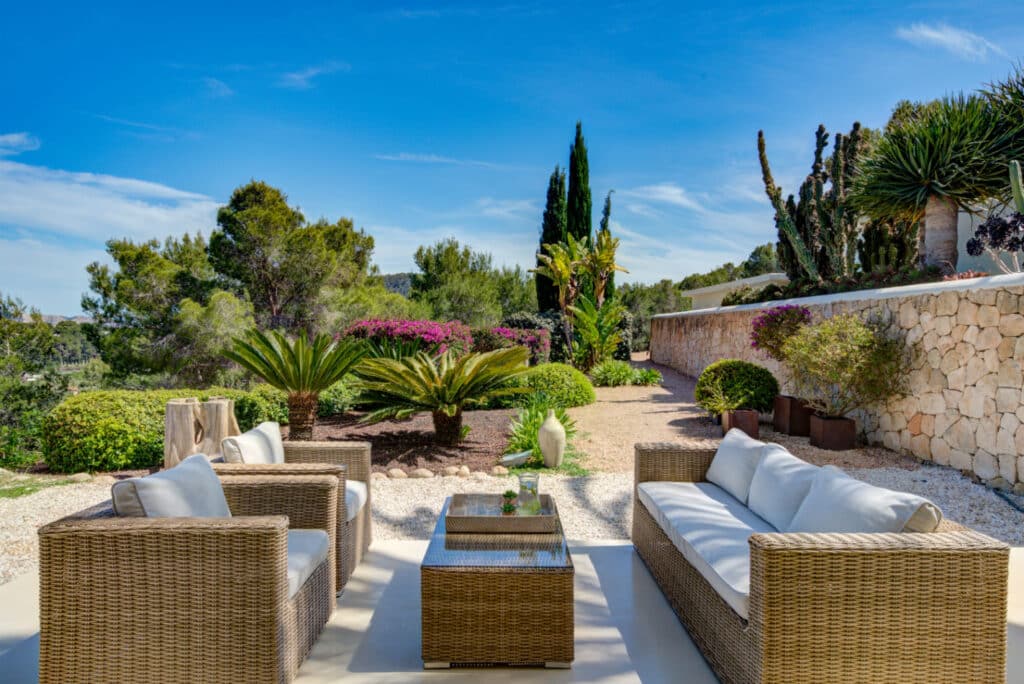 The outdoor seating area, perfect for luxury travel enthusiasts, boasts wicker sofas and a glass-top table on a patio, enveloped by lush greenery and a stone wall. The backdrop includes majestic trees set against clear blue skies. David Pearson Travel