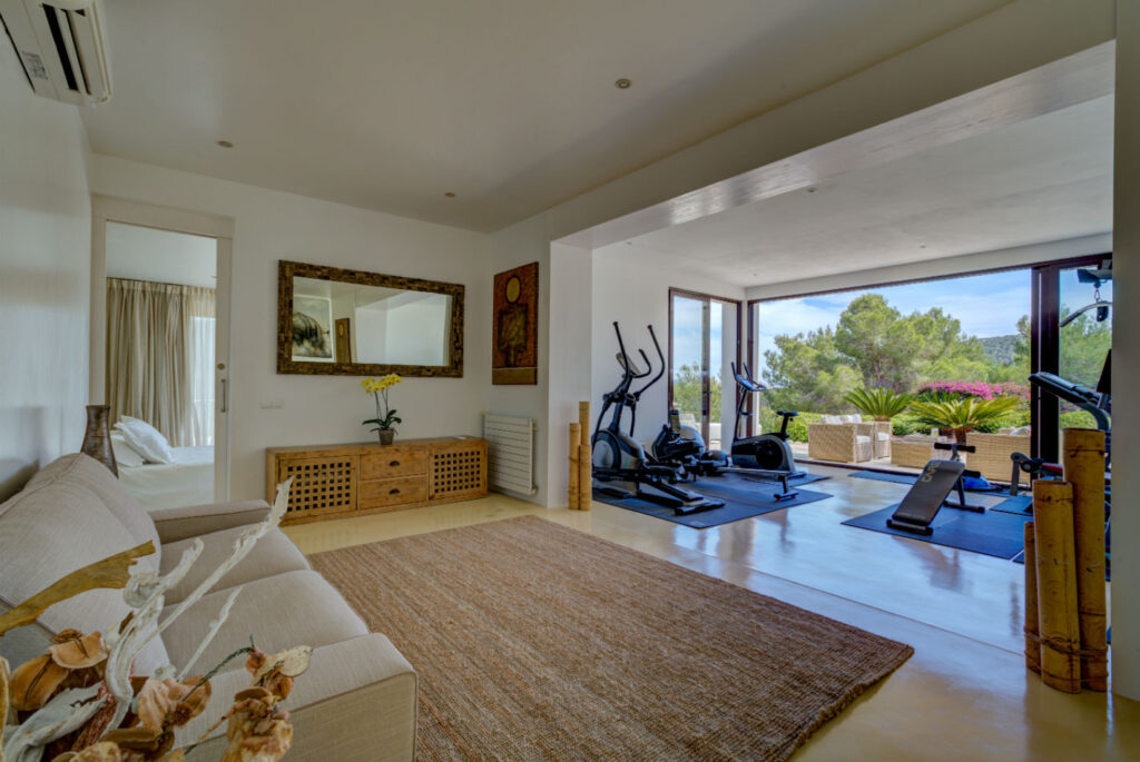 Spacious room with a seating area and home gym, perfect for luxury travel enthusiasts. A large window offers views of trees and a garden, flooding the space with natural light. The room features a rug, sofa, exercise equipment, and elegant decor elements like vases and a wall mirror. David Pearson Travel