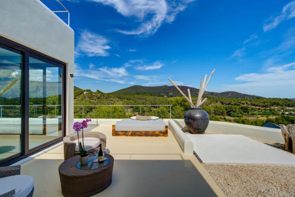 A modern terrace for luxury travel enthusiasts, featuring cushioned seating, wicker furniture, and a large decorative vase under a blue sky. It overlooks lush green hills and trees, with sliding glass doors reflecting the scenery. An orchid and a bottle with glasses sit elegantly on the table. David Pearson Travel