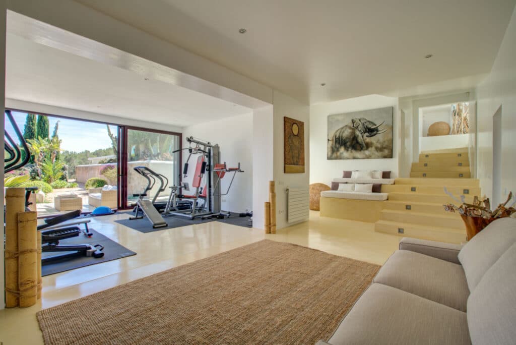 A bright, modern home gym with large windows opening to a garden exudes the allure of luxury travel. The room features exercise equipment like a treadmill and weight machine. A cozy seating area with a beige couch and a staircase adorned with artwork is visible. David Pearson Travel