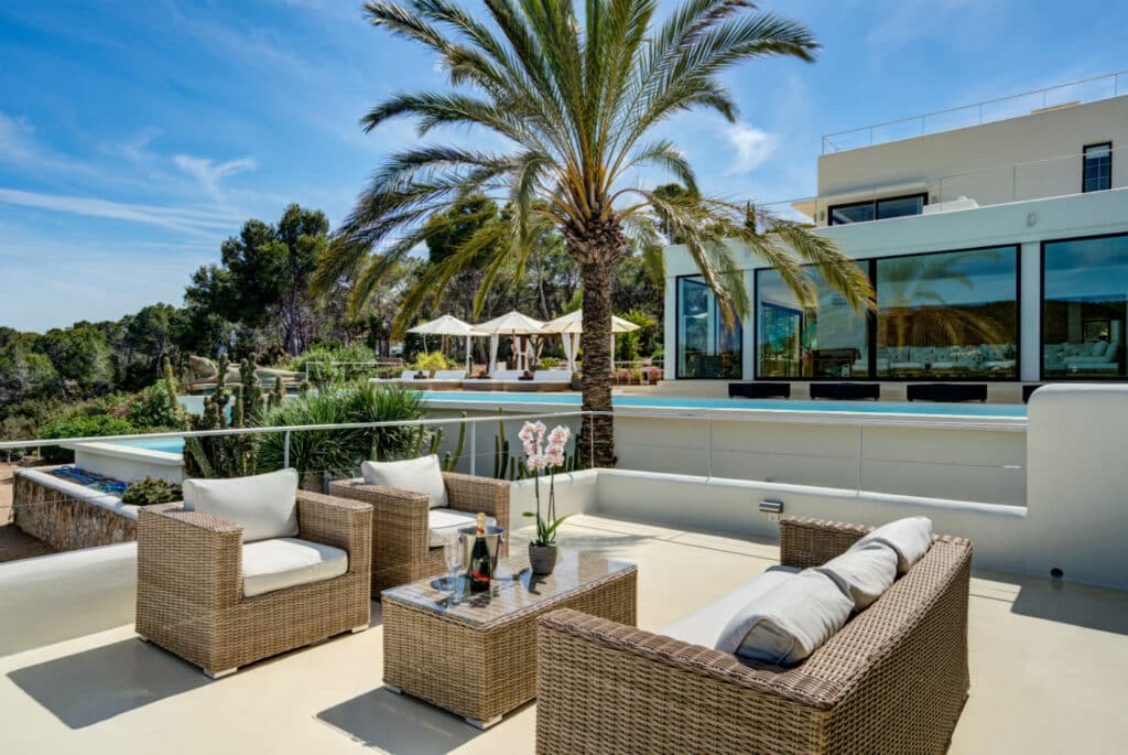 A luxurious outdoor patio, perfect for luxury travel enthusiasts, features wicker furniture with white cushions centered around a glass-top table. A palm tree provides shade, while a modern building with large windows offers views of lush greenery in the background. David Pearson Travel