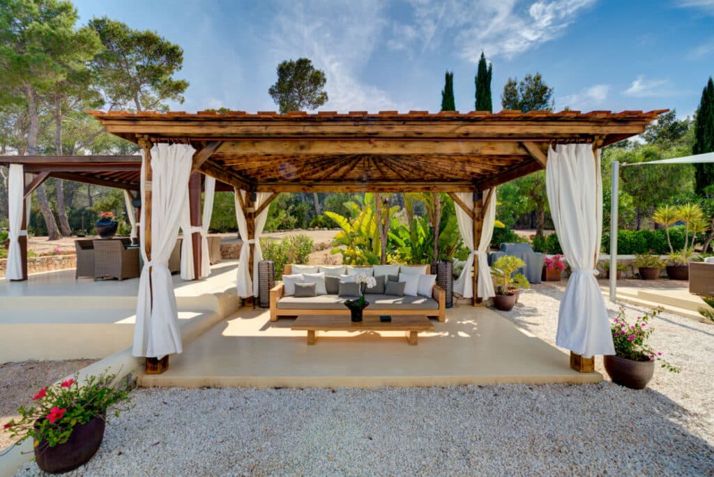 A wooden gazebo with a gabled roof and flowing white curtains is centered in the garden, offering a luxurious travel experience. Inside, cushioned seating and a coffee table rest on gravel, surrounded by potted plants and lush greenery under the expansive blue sky. David Pearson Travel