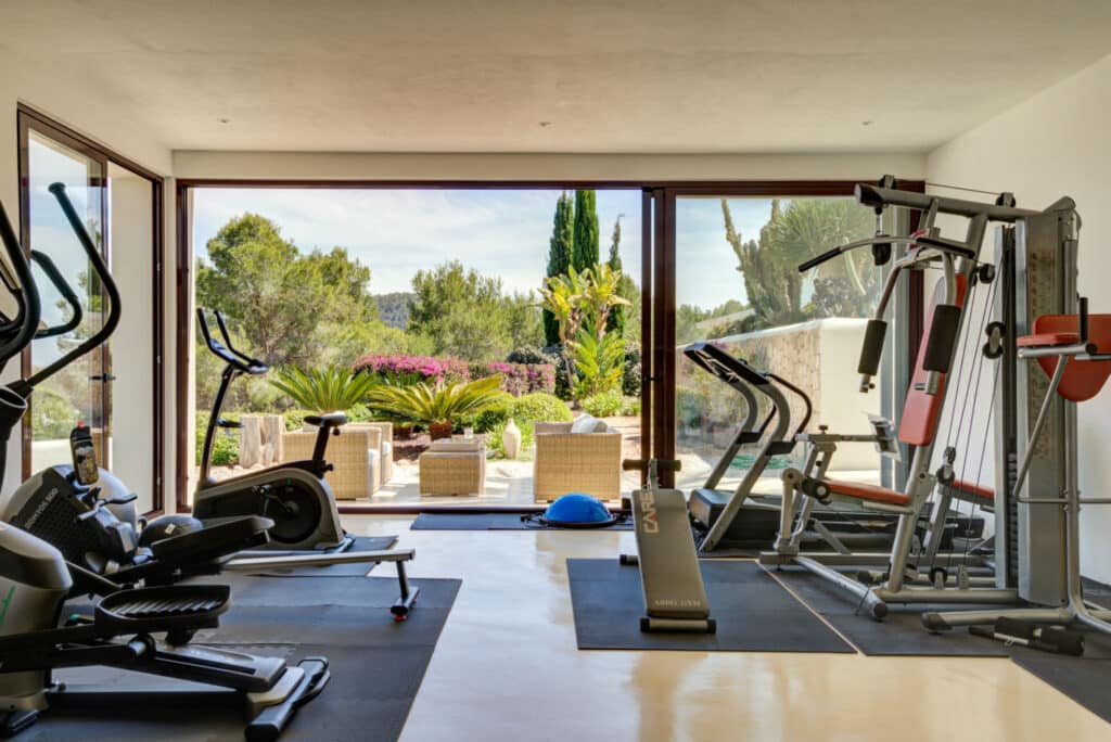 A home gym with luxury travel vibes features exercise equipment like a treadmill, elliptical, and weight machine, all facing large windows that reveal a lush garden brimming with various plants and trees. Elegant wicker furniture graces the patio outside. David Pearson Travel