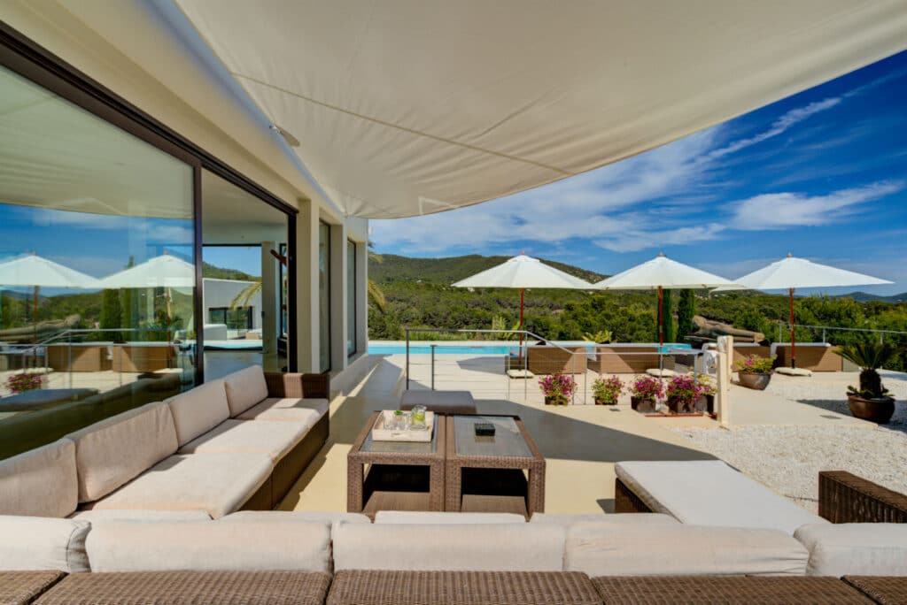 Experience the epitome of luxury travel on this exquisite outdoor terrace featuring wicker sofas and a wooden table. White umbrellas provide shade, while a glass railing reveals stunning views of the pool, lush green hills, and a blue sky. Potted plants enhance the areas natural charm. David Pearson Travel