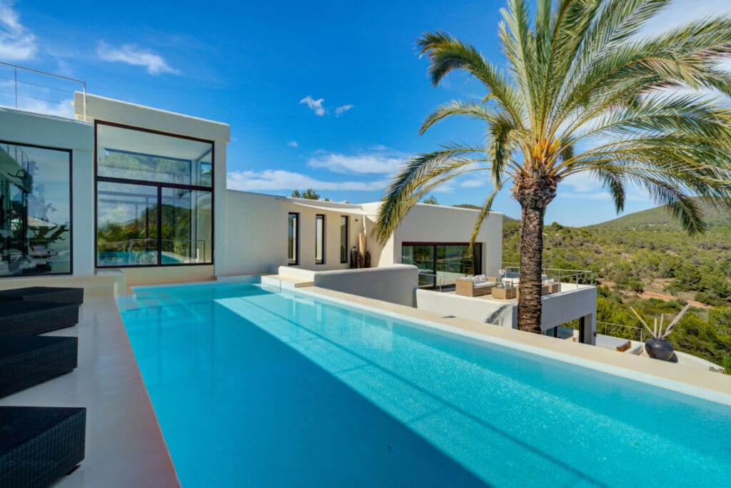 Experience ultimate luxury travel in a modern villa featuring a sleek infinity pool, surrounded by lounge chairs and overlooking lush greenery. Tall palm trees accentuate the contemporary architecture under a clear blue sky, offering an exquisite getaway. David Pearson Travel