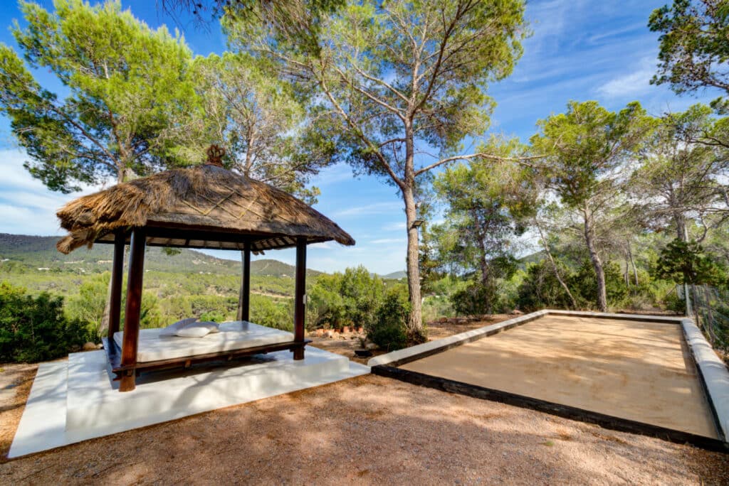Discover luxury travel at its finest in a serene outdoor area featuring a thatched-roof gazebo and lounge bed on a small platform, surrounded by pine trees. Nearby, enjoy a sandy petanque court with lush hills under a clear blue sky as your backdrop. David Pearson Travel