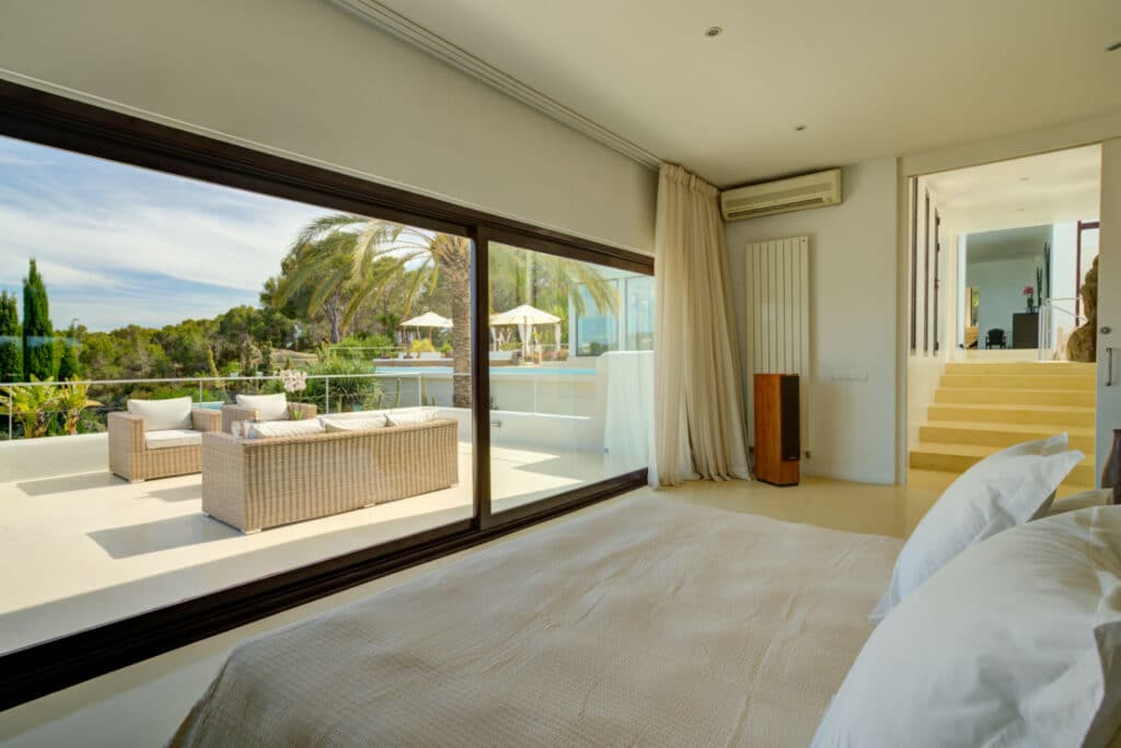 A bright bedroom epitomizing luxury travel features a large window overlooking a terrace. The terrace boasts wicker furniture with white cushions and views of palm trees and greenery. Inside, a bed with pristine white linens faces the window, while stairs ascend to another exquisite room. David Pearson Travel