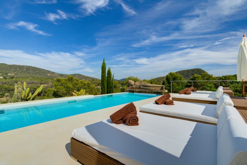 Luxurious outdoor pool area with cushioned loungers, rolled brown towels, and parasols under a blue sky. Lush green hills serve as a scenic backdrop, enhancing the serene and relaxing atmosphere—truly a haven for luxury travel enthusiasts. David Pearson Travel
