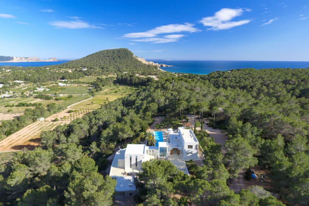 Aerial view of a modern white villa, epitomizing luxury travel, surrounded by dense greenery with a blue pool. Rolling hills and lush landscapes stretch towards the horizon. The distant blue sea and a partially cloudy sky complete the scenic backdrop. David Pearson Travel