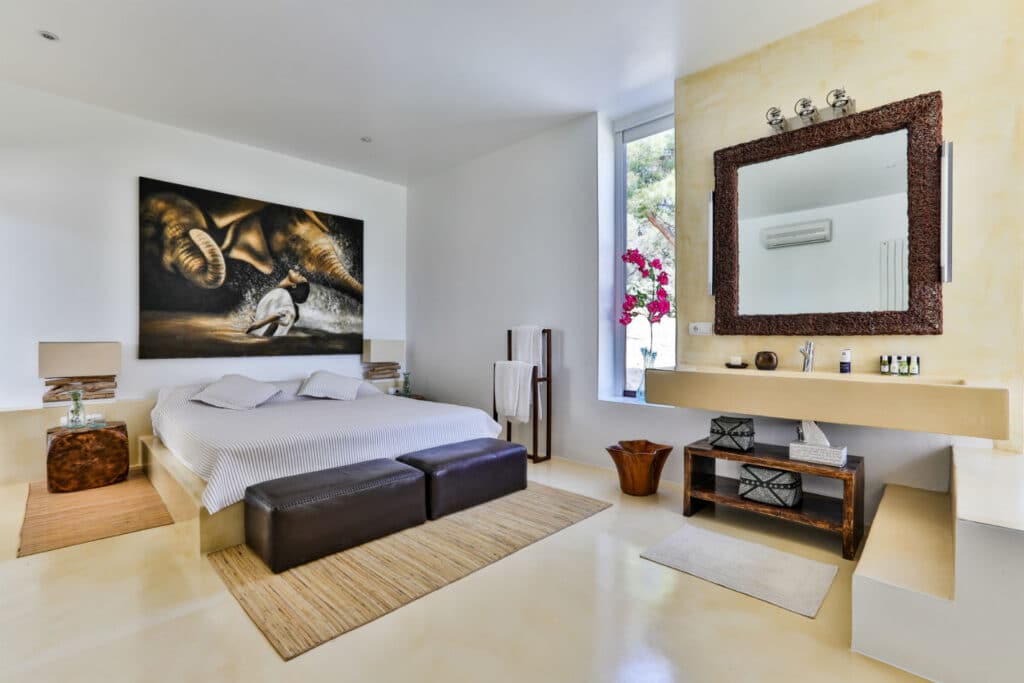 A modern bedroom exuding the elegance of luxury travel, with a large bed, two dark benches at the foot, and a stunning painting above. A rustic vanity with a mirror and pink flowers sits beside a tall window. The rooms minimalistic decor in neutral tones is bathed in natural light. David Pearson Travel