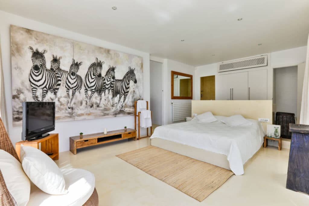 A modern bedroom perfect for luxury travel enthusiasts features a large bed with white linens and a zebra-themed art piece. A flat-screen TV on a wooden stand complements the woven seating, while minimalistic decor in neutral tones is bathed in natural light. David Pearson Travel