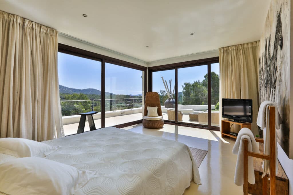 A bright bedroom with large windows overlooking a scenic view of hills and trees offers a touch of luxury travel. The room features a bed with white linens, a wooden chair, a small TV, and outdoor seating on the balcony. Neutral-toned curtains gracefully frame the windows. David Pearson Travel