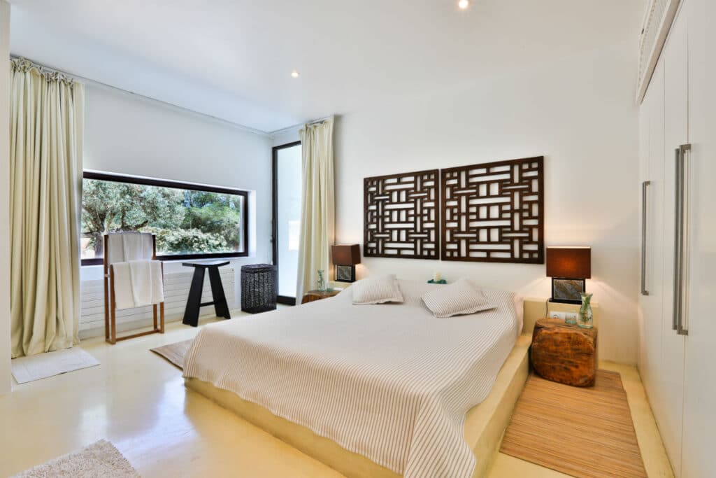 A modern bedroom exuding luxury travel vibes, with a large bed featuring neutral-colored bedding. Decorative panel above the headboard, two side tables with lamps, and a plush rug underneath. A large window on the left floods in natural light. Minimalist decor and light walls complete this serene retreat. David Pearson Travel