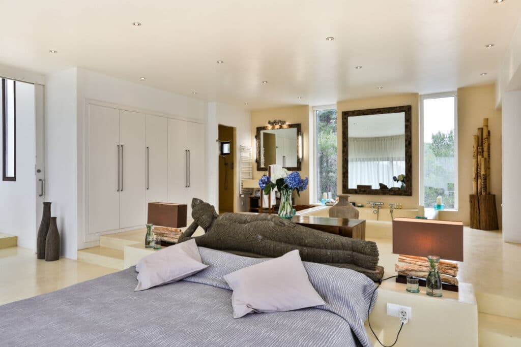 Spacious bedroom with a large bed featuring pillows and a striped duvet, flanked by two rustic lamps. A mirror, flowers, and decor items are in the background. Bright natural light fills the room through wide windows, offering a touch of luxury travel ambiance. David Pearson Travel