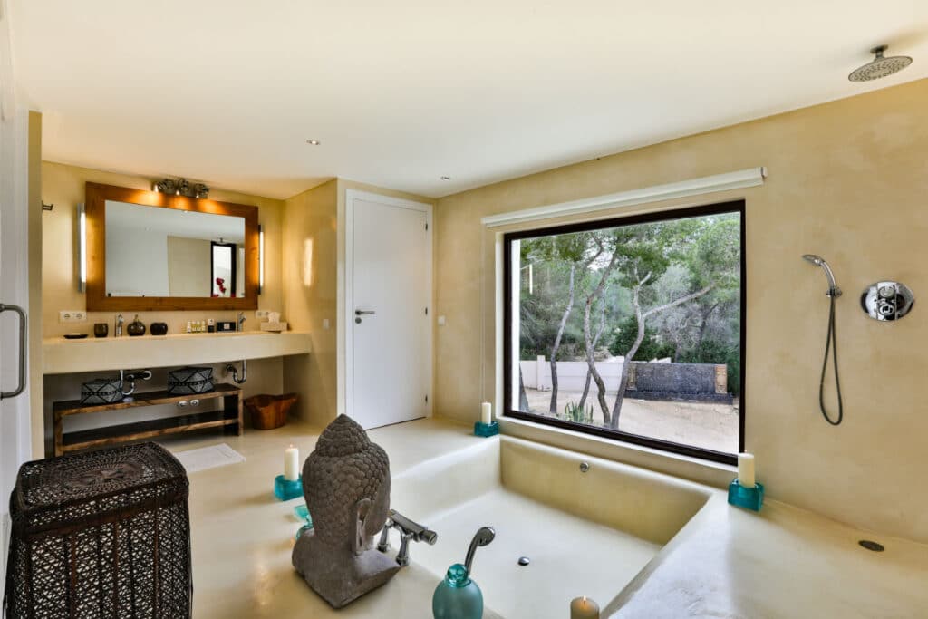 A modern bathroom reminiscent of luxury travel, featuring a sunken bathtub, a large window with tree views, and a wooden vanity with a mirror. Decorative elements like candles and a sculpture enhance the cream-colored walls, crafting an ambiance of serene elegance. David Pearson Travel