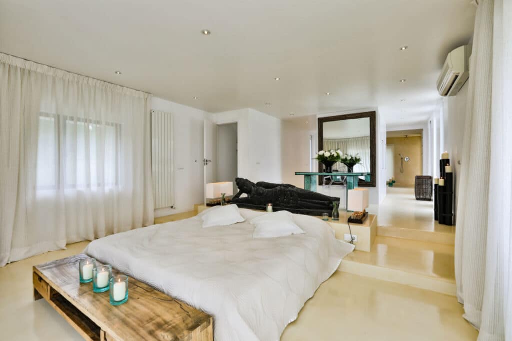 The minimalist bedroom exudes an air of luxury travel with its low wooden platform bed, pristine white bedding, and decorative candles. A large statue stands by a grand mirror. Soft lighting bathes the room, complemented by sheer curtains and an air conditioner. A corridor invites exploration to another area. David Pearson Travel