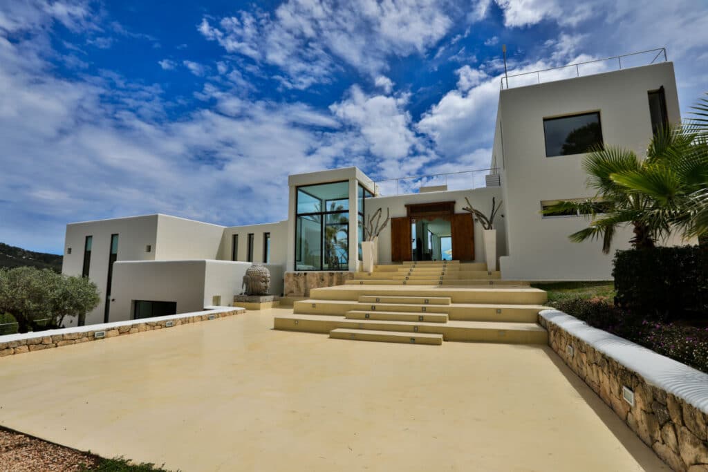 Experience luxury travel at its finest in this modern white villa with large windows, a flat roof, and a grand entrance featuring wide stone steps. Surrounded by palm trees and lush greenery, it sits under a clear blue sky adorned with scattered clouds. David Pearson Travel
