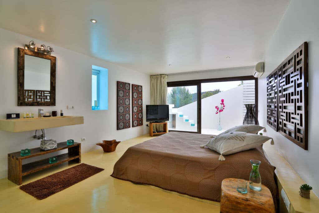 Modern bedroom with a large bed covered in a brown quilt, perfect for a touch of luxury travel at home. Features include a wooden sink, decorative wall panels, TV, and glass doors to an outdoor area. Minimalist decor with white walls and ceiling creates a bright, airy feel. David Pearson Travel