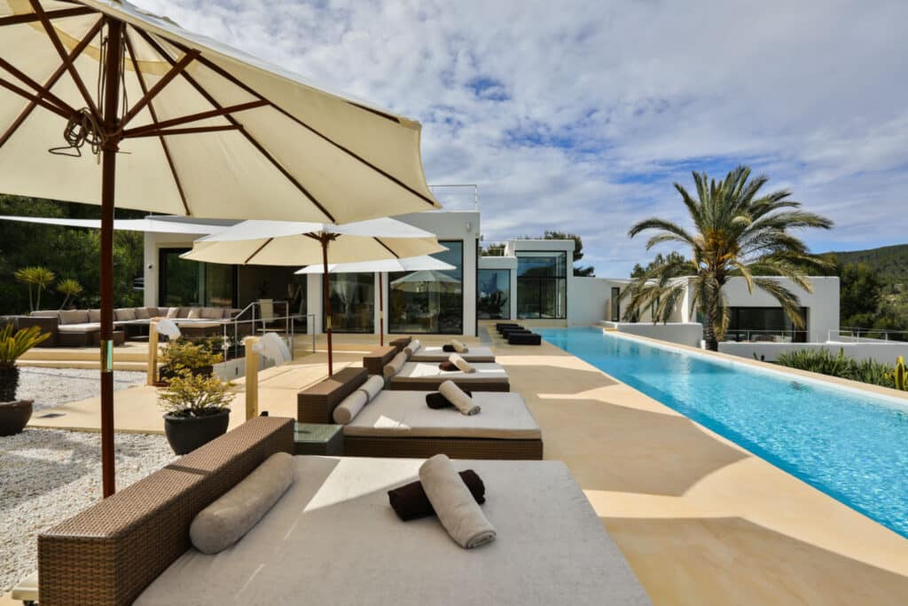 Experience luxury travel at its finest in this outdoor oasis, complete with modern sun loungers under large umbrellas. A sleek rectangular pool, surrounded by palm trees and contemporary architecture, invites relaxation beneath a partly cloudy sky. David Pearson Travel