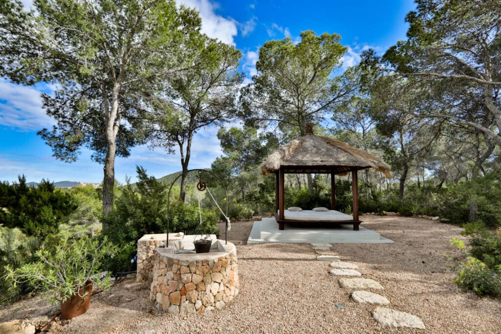 A secluded hammock bed under a straw-roofed gazebo offers a luxury travel experience, surrounded by tall trees and lush greenery. A stone path leads to a rustic stone well, all beneath a clear blue sky, perfect for unwinding in natures embrace. David Pearson Travel