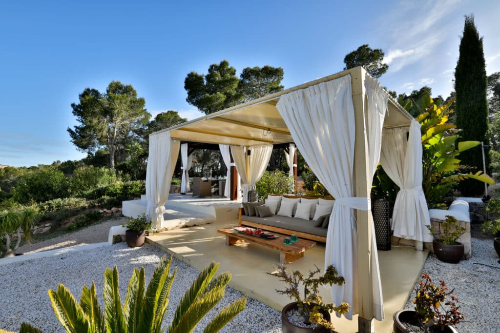 Nestled in the garden, an outdoor canopy with white curtains exudes a sense of luxury travel. Beneath, a wooden table and cushioned seating invite relaxation. Surrounded by potted plants and lush greenery under a clear sky, it creates an exquisitely serene atmosphere. David Pearson Travel