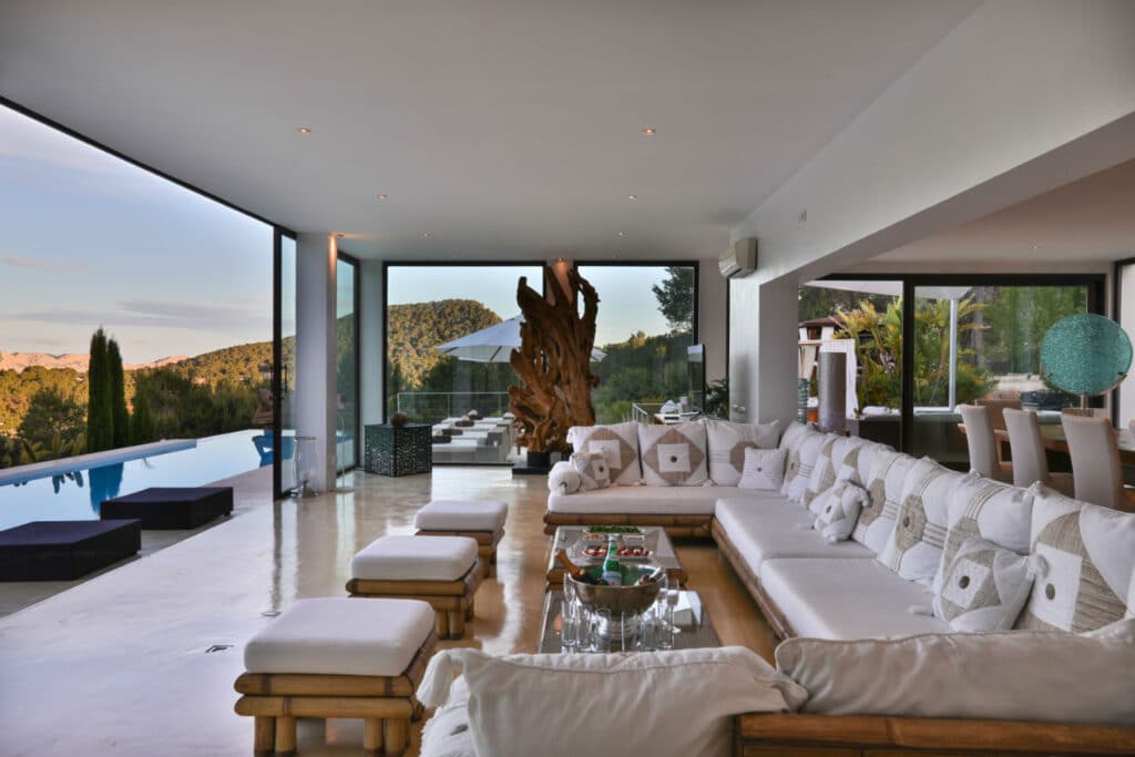 A haven of luxury travel vibes, this living room boasts a large white sectional sofa and wooden furniture. Floor-to-ceiling windows reveal an outdoor pool against scenic hills. A glass coffee table and decorative sculpture complete the scene, with a dining area subtly in view. David Pearson Travel