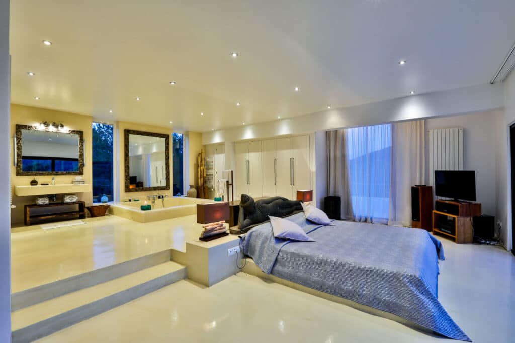 Experience luxury travel in this spacious, modern bedroom featuring a large bed and cozy seating area. Enjoy the sleek decor with a luxurious bathroom, ambient lighting, and sheer-curtained windows. The minimalist design exudes elegance and offers a serene retreat. David Pearson Travel