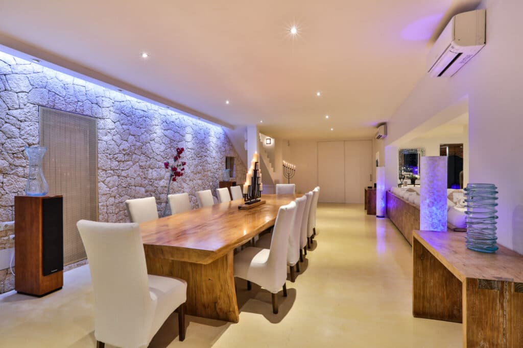 Luxurious dining room with a long wooden table and white upholstered chairs, reminiscent of an upscale luxury travel experience. Stone accent wall with soft blue lighting and mirrors add to the modern, ambient feel. Decorative elements enhance this polished, sleek design. David Pearson Travel
