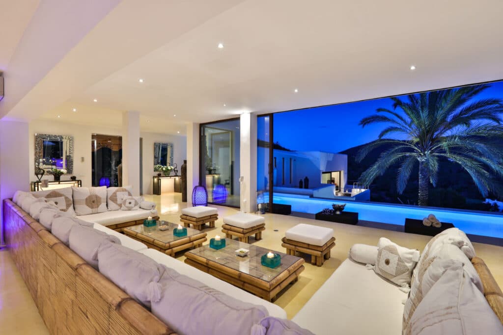 Experience luxury travel with this exquisite open-air living space featuring white sofas and a wooden coffee table, all overlooking an illuminated swimming pool. A large palm tree and distant hills enhance the view, framed by expansive glass doors under the deep blue evening sky. David Pearson Travel