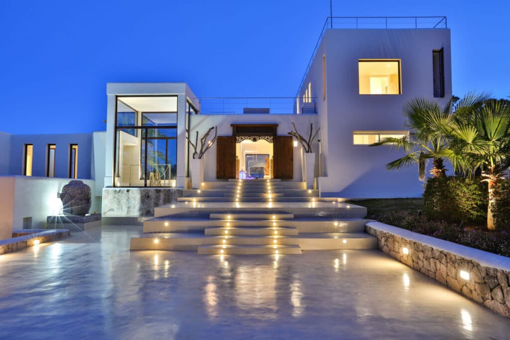 At dusk, the modern villa exudes luxury travel vibes with its glowing pathway lights guiding you to a grand entrance. Large windows, minimalist design, and landscaped greenery, including palm trees, accentuate the serene atmosphere against a deep blue sky. David Pearson Travel