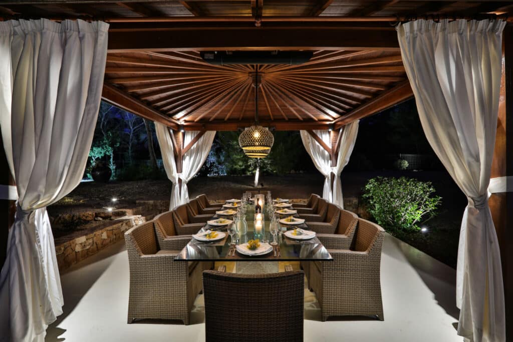 The luxury travel experience extends to the outdoor dining area under a wooden gazebo, where a long table awaits surrounded by wicker chairs. A warm chandelier adds elegance, while white curtains and softly lit greenery create an inviting ambiance. David Pearson Travel