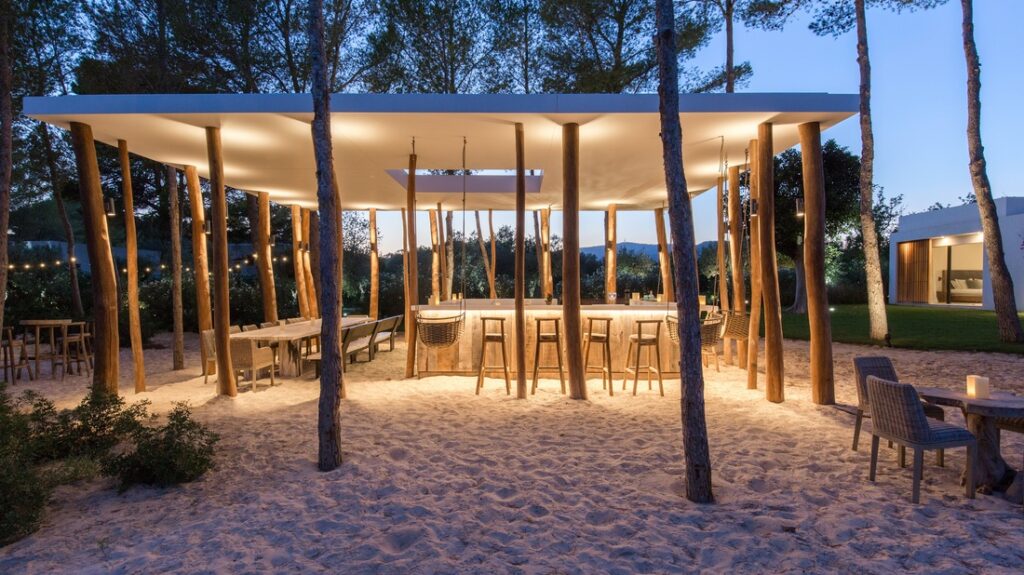 An outdoor bar with a modern design, supported by uneven wooden posts. Situated on sand and surrounded by trees, it offers an exclusive luxury travel experience. Bar stools and tables are elegantly arranged, and the area is softly lit, providing a cozy evening ambiance. David Pearson Travel