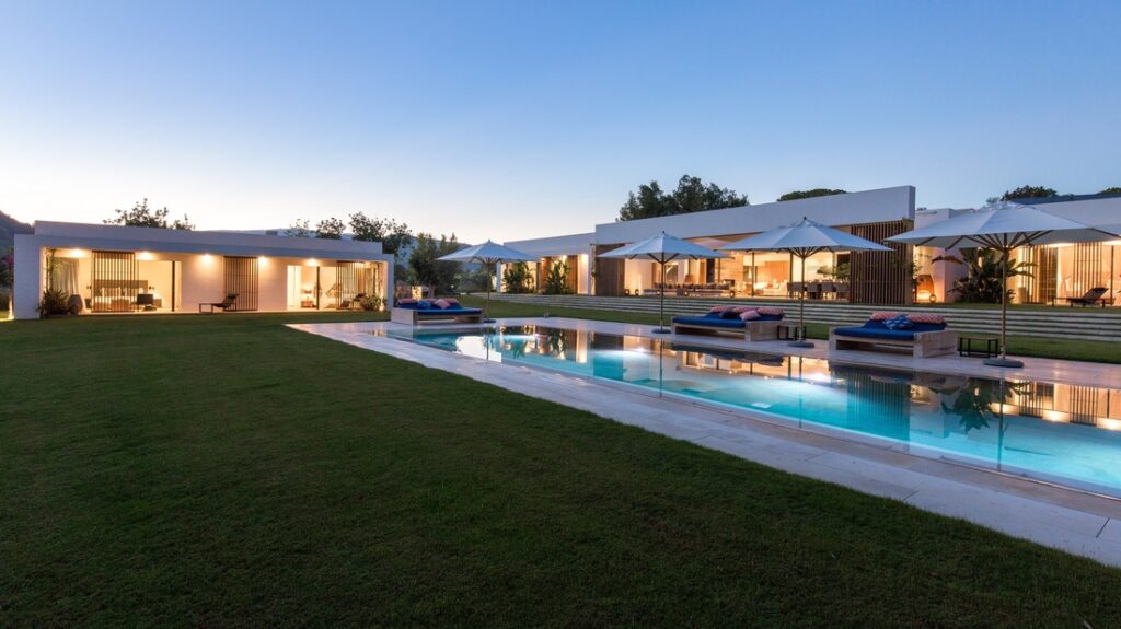 Experience the epitome of luxury travel in this modern house, featuring large glass windows and an inviting outdoor pool. Nestled amidst a well-manicured lawn, sun loungers with cushions and umbrellas await you by the pool as the house glows warmly in the evening light. David Pearson Travel