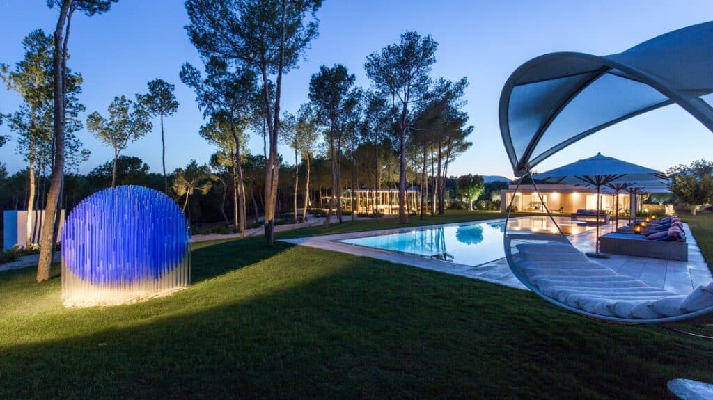 A modern outdoor setting at dusk exudes luxury travel vibes, featuring a swimming pool with lounge chairs and umbrellas. A spherical, illuminated blue sculpture graces the lawn, surrounded by tall trees, creating a tranquil and elegant atmosphere. David Pearson Travel