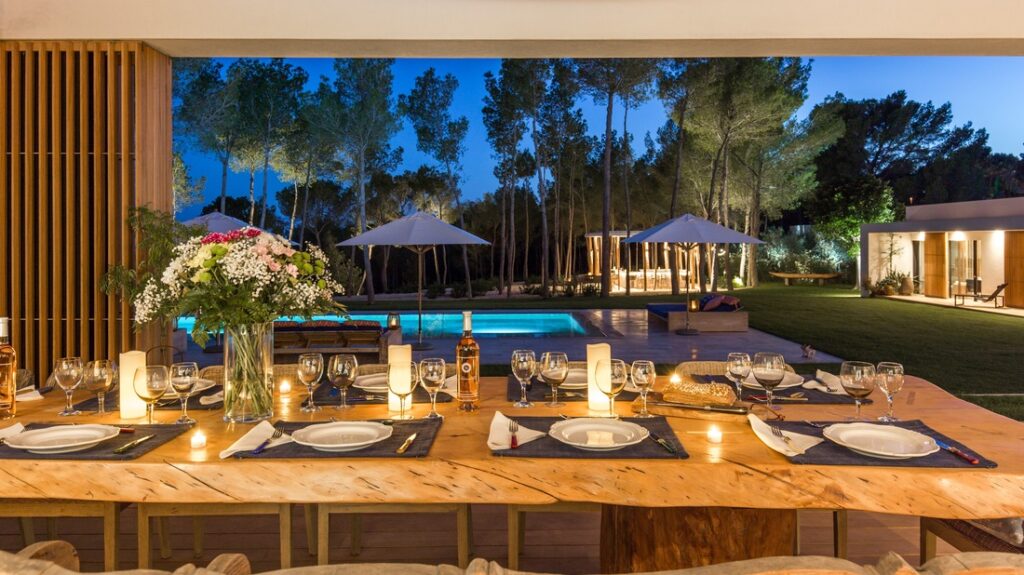A beautifully set outdoor dining table with candles and flowers overlooks a pool adorned with lit umbrellas and trees in the background, epitomizing luxury travel. The scene at dusk is illuminated by warm lighting from nearby structures, enveloped in a tranquil garden ambiance. David Pearson Travel