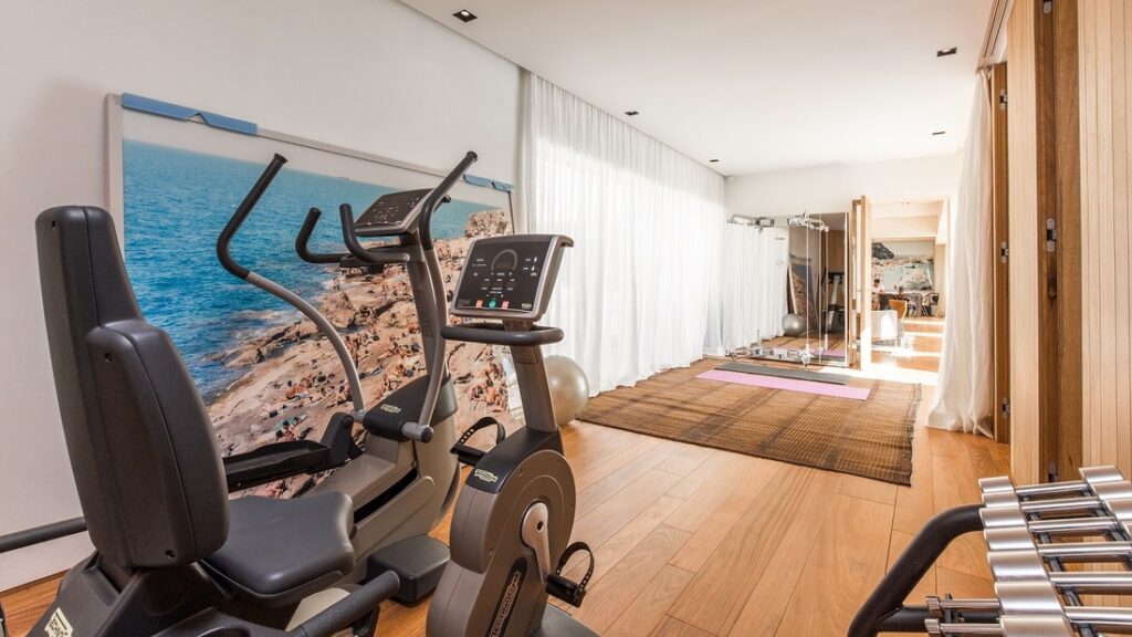 A home gym designed for luxury travel enthusiasts, featuring exercise equipment like an elliptical machine and squat rack. The room boasts wood flooring, large windows with sheer curtains, and a beach-themed wall mural. Dumbbells are neatly displayed on a rack to the right. David Pearson Travel