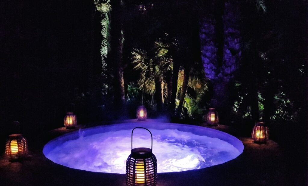 A round hot tub with glowing purple water, ideal for luxury travel enthusiasts, is surrounded by lit lanterns at night. Palm trees and foliage form a dark backdrop, creating a tranquil and inviting atmosphere. David Pearson Travel