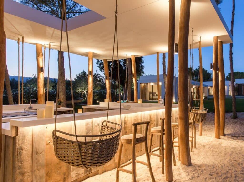 Outdoor bar with wooden stools and swing seats under a canopy, surrounded by tall tree trunks. The warmly lit area sits on a sandy surface, creating a relaxed atmosphere reminiscent of luxury travel. In the background, a modern building is partially visible. David Pearson Travel