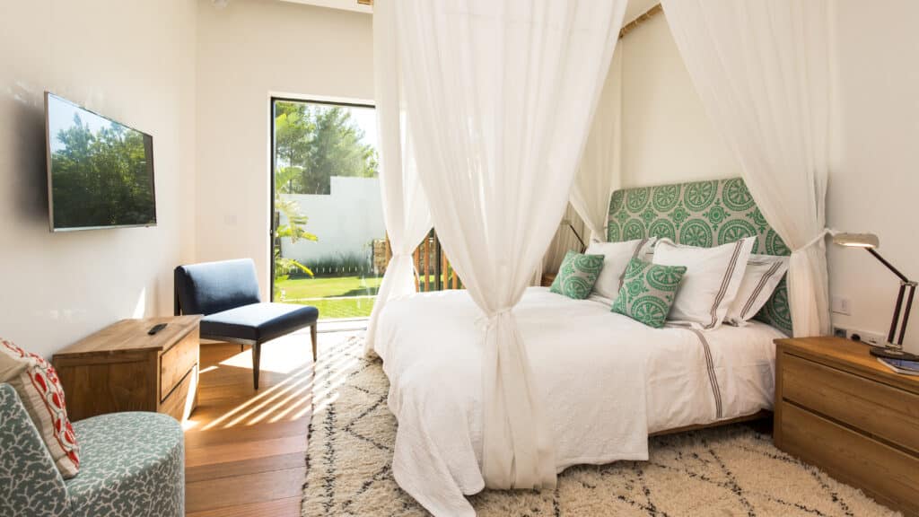 Bright bedroom with a luxurious four-poster bed draped in white curtains and green-patterned pillows. A blue chair sits by the large window, offering a travelers view of the garden. Wooden nightstands flanking the bed hold a lamp and decor, while a wall-mounted TV is on the left. David Pearson Travel