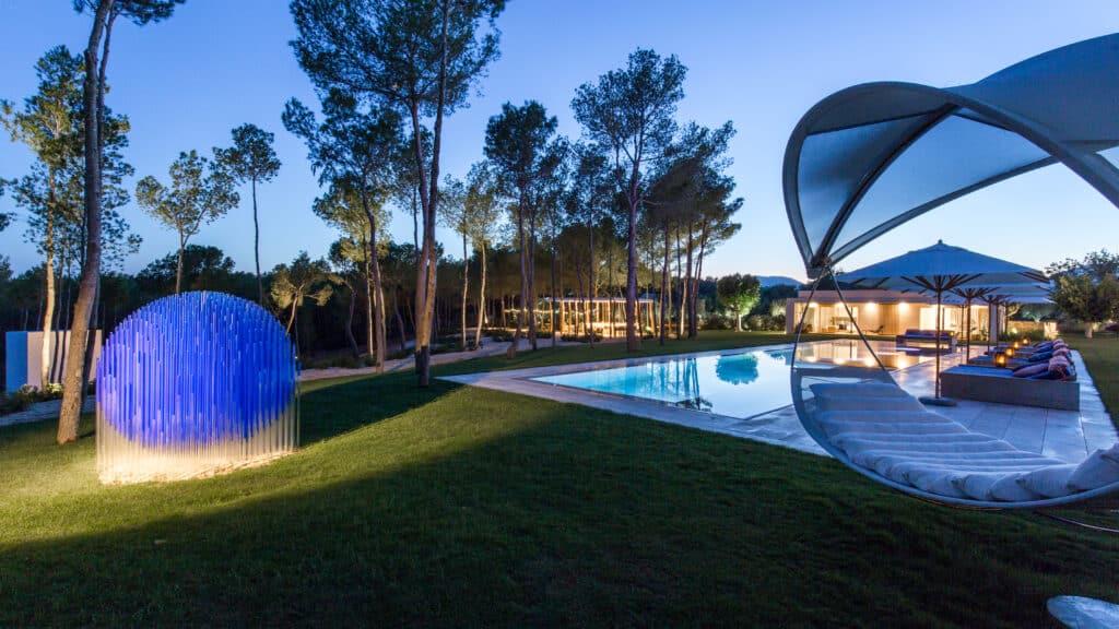 A modern outdoor setting at dusk exudes the essence of luxury travel, featuring a swimming pool, lounge area with a canopy, and an illuminated blue spherical sculpture. Trees surround the area as the sky elegantly transitions to night. David Pearson Travel