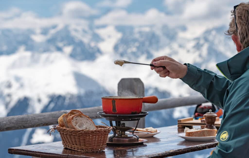 A person enjoys cheese fondue outdoors with a view of snow-covered mountains—one of the top things to do in Verbier—dipping bread into a red pot on a wooden table set with bread, a basket, and condiments. David Pearson Travel