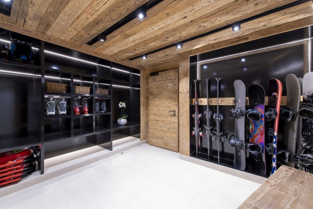 A modern ski storage room with wooden walls and ceiling, black shelves holding helmets and boots, and racks displaying skis and snowboards. The room is brightly lit with recessed ceiling lights. David Pearson Travel