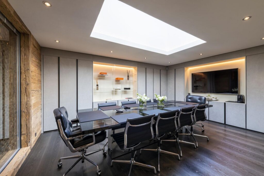 Modern conference room with a large black glass table surrounded by eight black chairs, skylight above, built-in shelves, a large screen on the wall, and vases of white flowers on the table. David Pearson Travel
