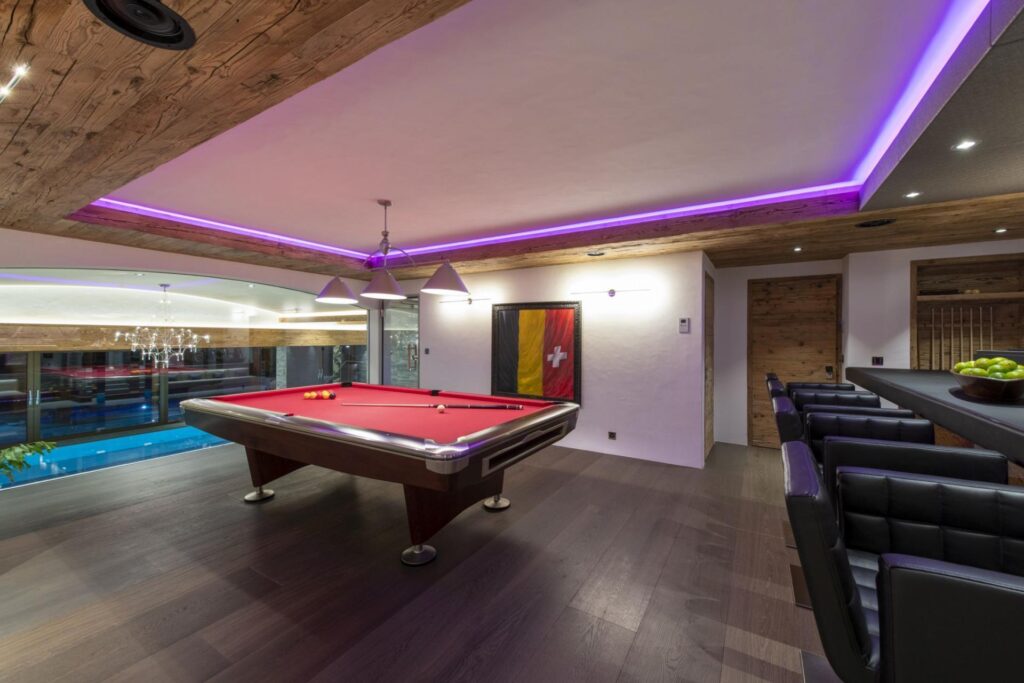 Modern game room with a red pool table, bar seating, and wood accents. Purple LED lighting illuminates the ceiling. A window overlooks an indoor pool, and a Swiss flag artwork hangs on the wall. David Pearson Travel