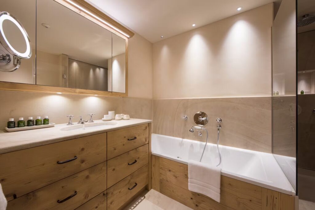 A modern bathroom with wooden cabinets, a double sink, a large mirror, a built-in bathtub, soft lighting, and neatly arranged toiletries. A white towel hangs over the edge of the tub. David Pearson Travel